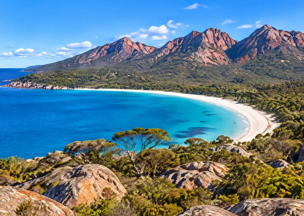 Panoramic elevated view of crescent white sand beach and turquoise ocean water framed by The Hazards granite peaks in Freycinet Tasmania